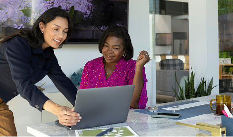 Two professionals collaborate in an office; one is standing while showing their laptop screen to a colleague at a desk.
