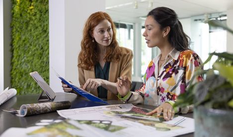 Two women sitting at a desk reviewing printed design plans in an office.