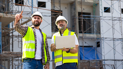 A project owner points at a tablet while speaking with a construction worker at a construction site.