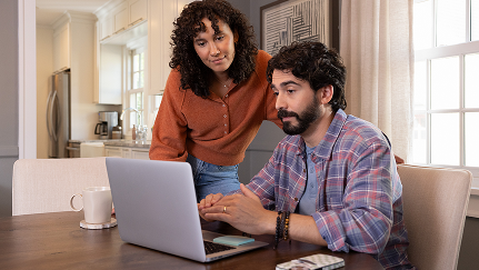 A middle aged married couple gathers in their dining room to read about annuities on their laptop.