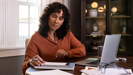 A middle aged woman takes notes and conducts research on her laptop while sitting at her dining room table.