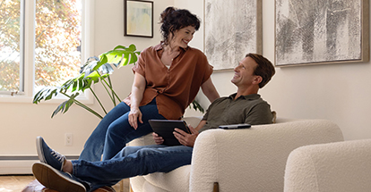 A middle-aged couple sits in an oversized chair smiling at each other. The husband holds a tablet while the wife sits on the arm of the chair.