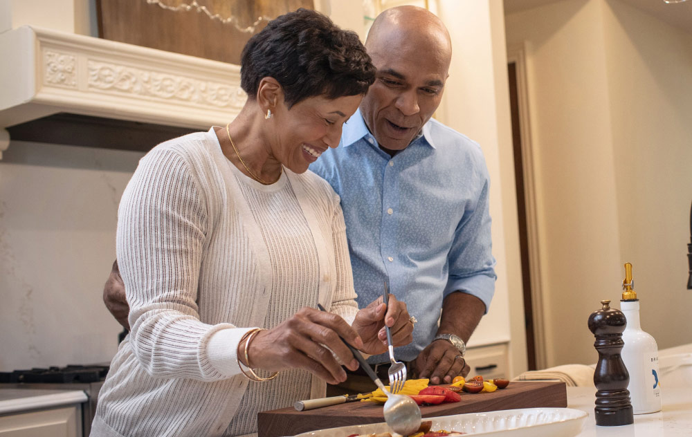 A middle aged married couple laughs while preparing dinner together in their kitchen.