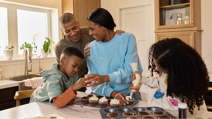 A family of four laughs as they decorate cupcakes together in their kitchen.