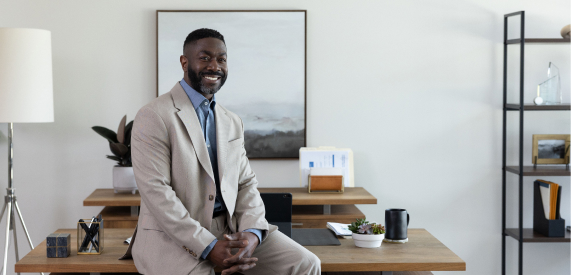 A well-dressed and smiling advisor leans confidently on the edge of his desk.