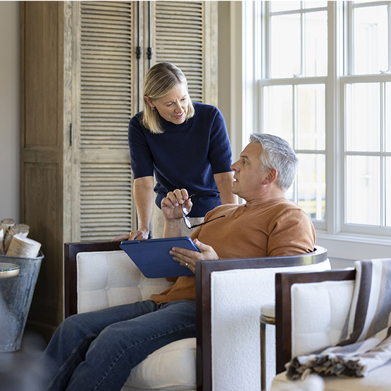 Middled-aged married couple discuss their personalized retirement income strategy in their living room. The husband shares insights from his tablet device with his wife.